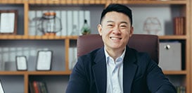 A smiling businessman sitting comfortably at his desk in front of a wooden bookshelf.