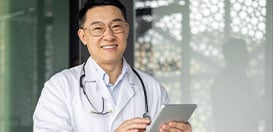 A smiling male doctor in a white coat and stethoscope holding a digital tablet.