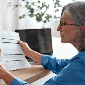 Woman wearing glasses and a blue shirt holding documents while sitting at a desk with an open laptop in a modern office setting.