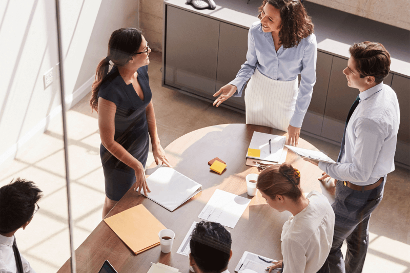 A group of colleagues stands around a table in a meeting at the office, discussing enterprise IT asset lifecycle management strategies, including ITAD, with a focus on degaussing services, IT asset remarketing,redeployment.