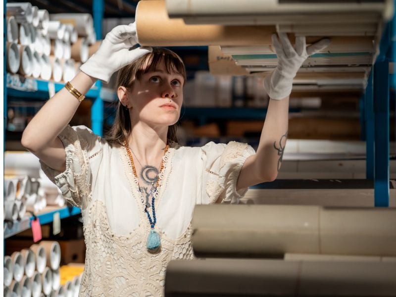 Person wearing white gloves carefully places or retrieves cardboard tubes on a shelf in a storage archive filled with rolled materials.