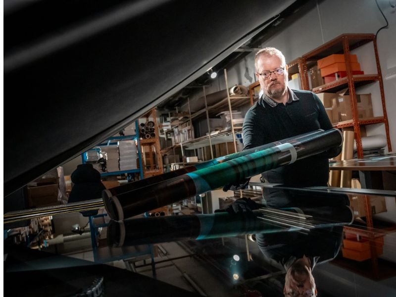 Man with glasses and gloves holding a rolled print above a reflective flatbed surface in a workshop setting.