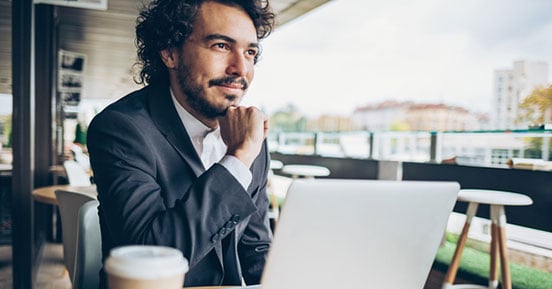 man sitting outside at a cafe with his laptop smiling and staring off