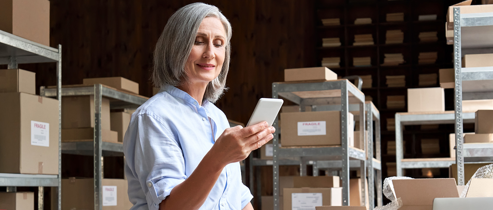 an older woman looking at phone in logistics backroom with packages
