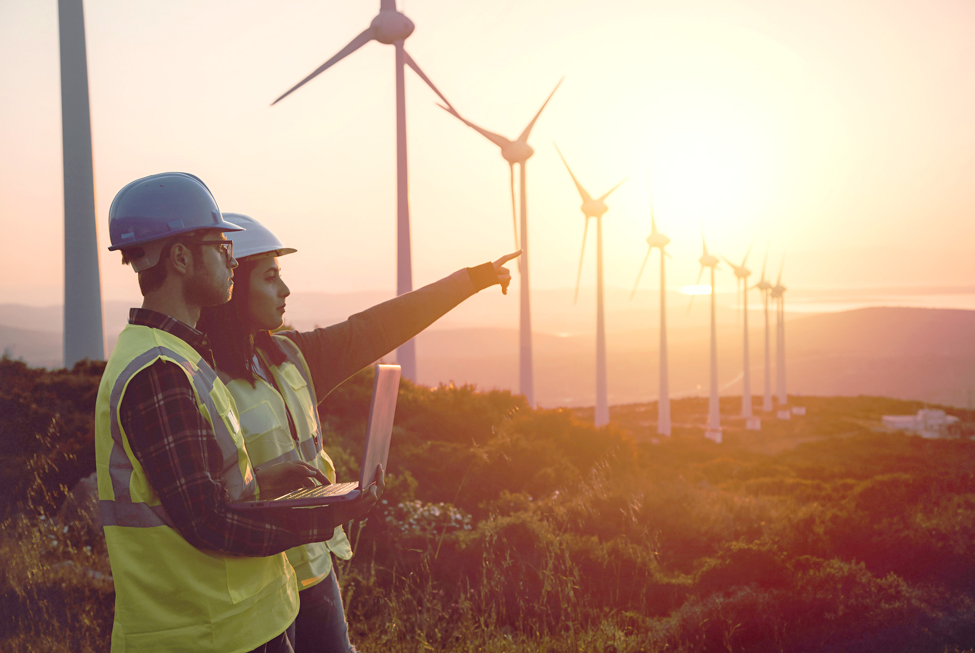 two energy workers pointing at windmills during sunset