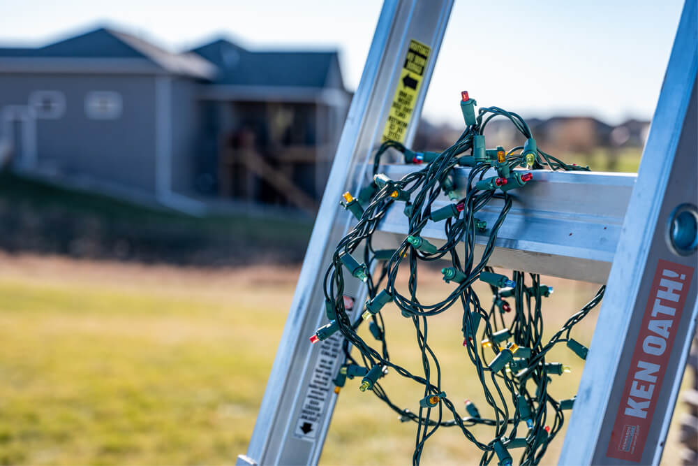 Close-up image of a ladder with Christmas lights on it, a house blurred in the background