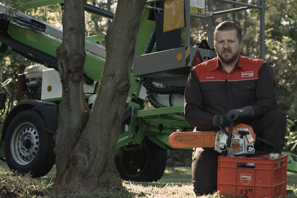 A Kennards Hire employee kneeling next to a chainsaw and a tree 