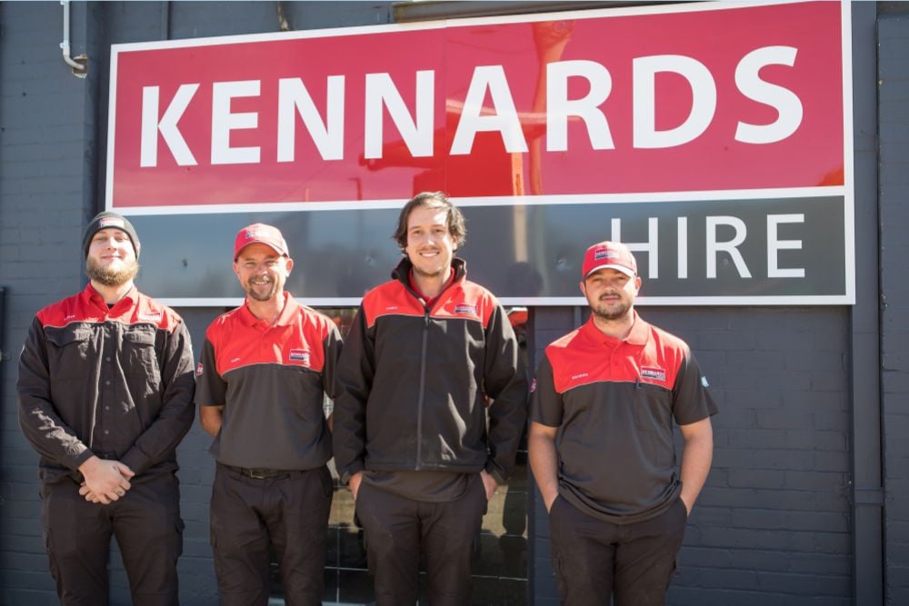 Four Kennards employees standing in front of a large Kennards Hire logo