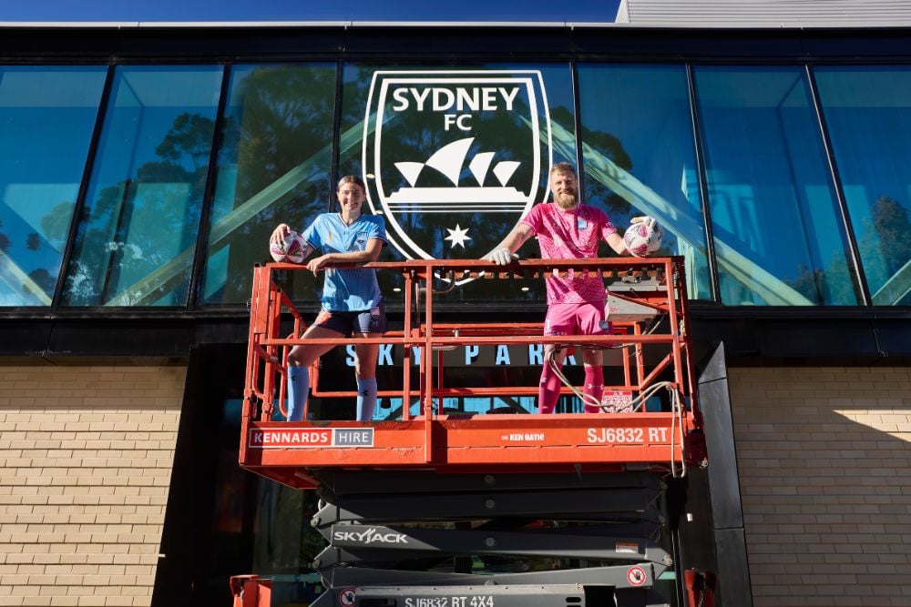 Sydney FC soccer players standing in a scissor lift in front of the Sydney FC training facility