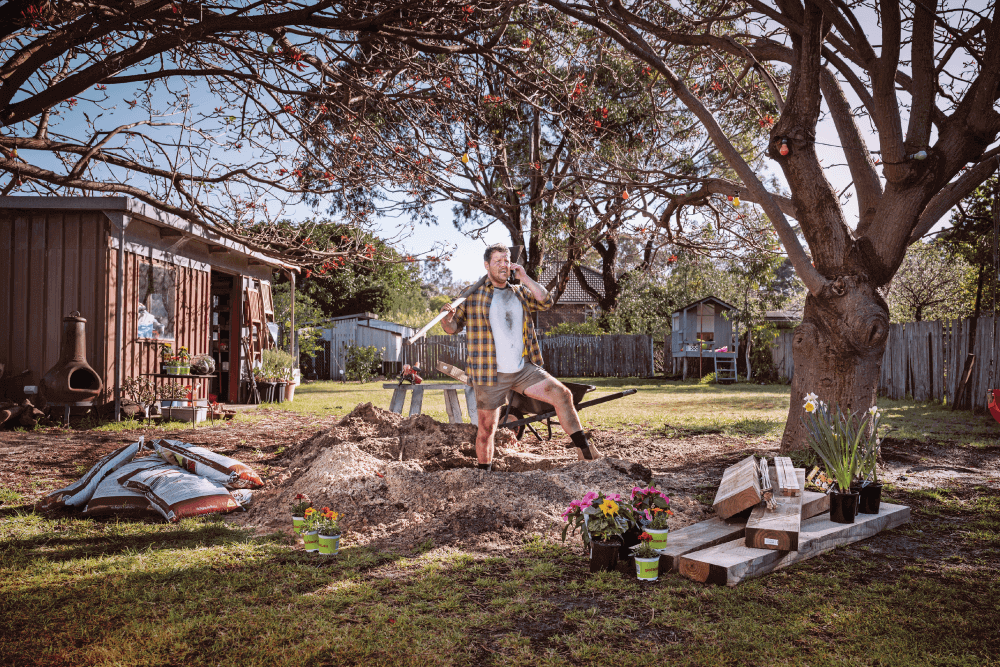 A DIYer standing in the middle of a backyard on the phone, shovel in hand