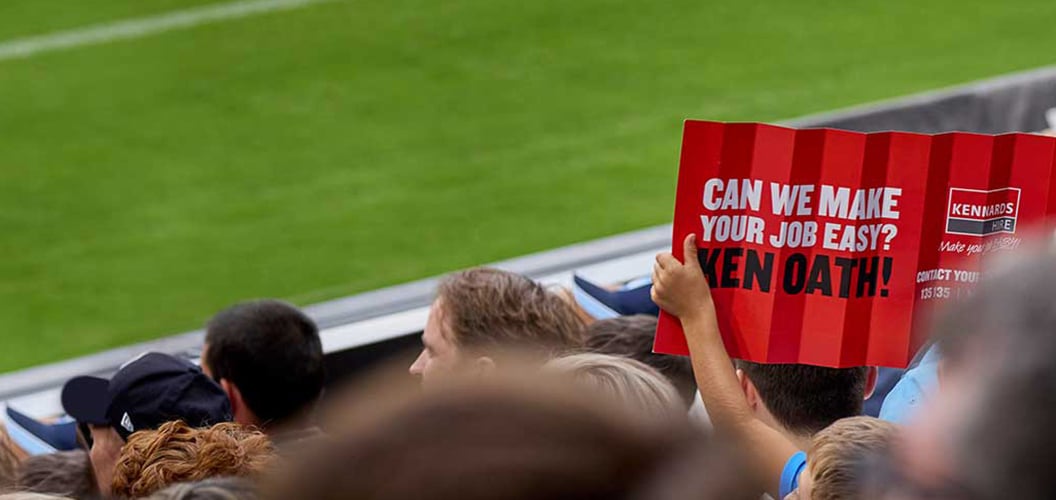 Picture of fans from behind looking onwards at a football field, a Kennards Hire  sign being held up