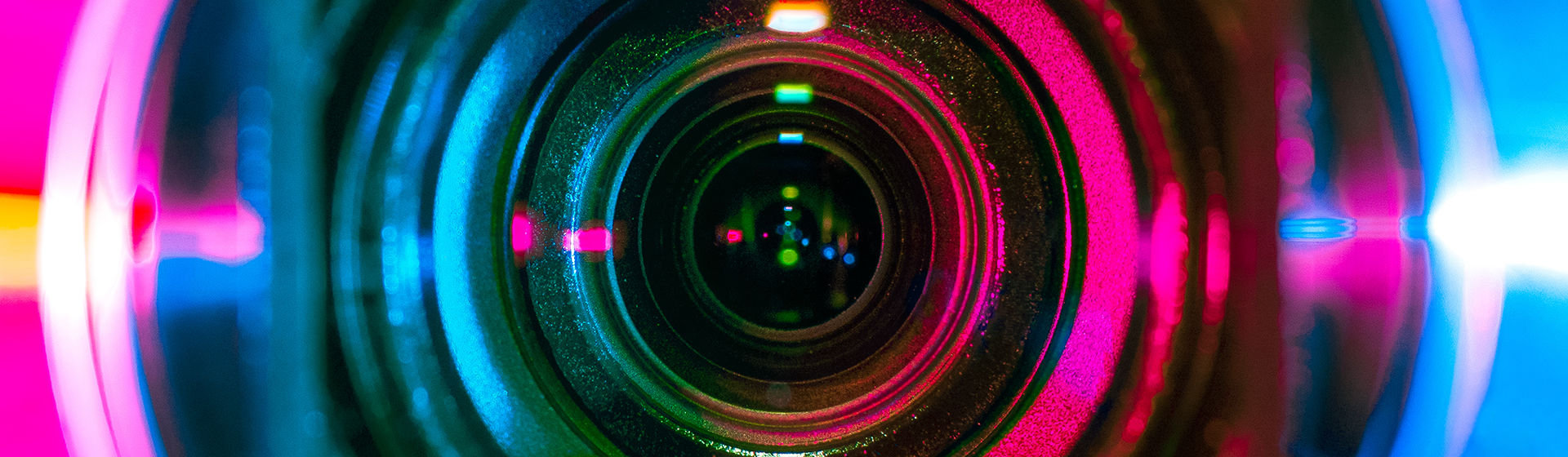 Lewis Silkin Close up camera lens in sharp focus, lit from the left in vibrant pink and the right in blue with shades of green in the lens