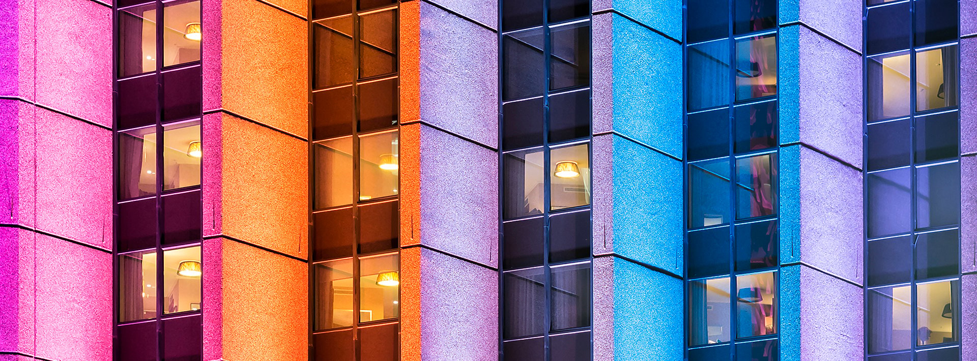 Lewis Silkin Close up of apartment building windows, the building exterior is lit in vertical strips in pink, orange, purple & blue light