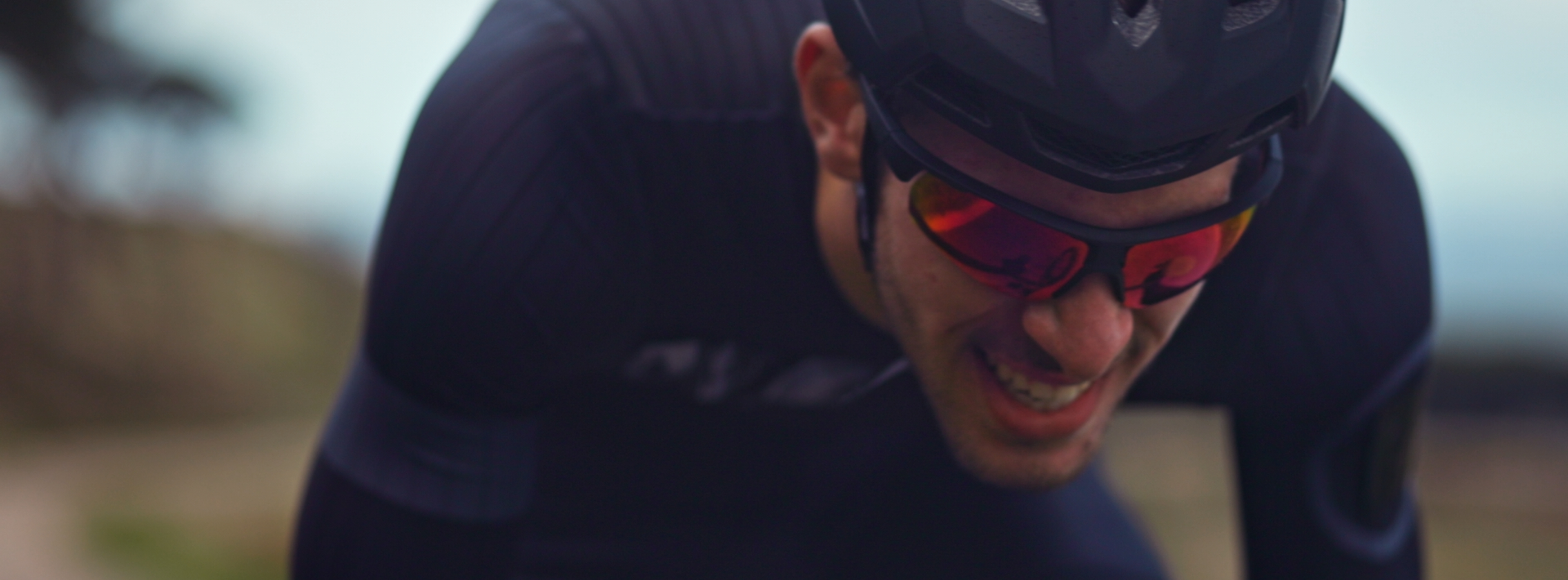 Close up of a cyclist in a dark blue bodysuit, helmet and red glasses