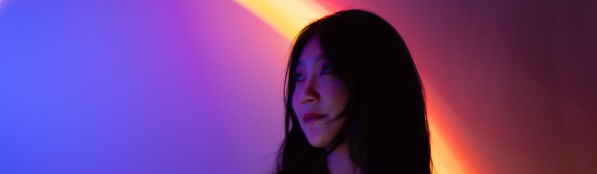 Starkly lit person with bright blue & purple light from the left, behind is a ring of orange light