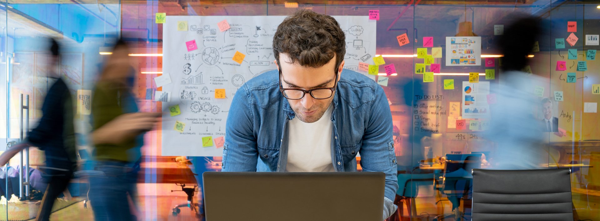 Lewis Silkin Person using a laptop in a meeting space with several figures walking through. Behind is a glass wall covered in diagrams