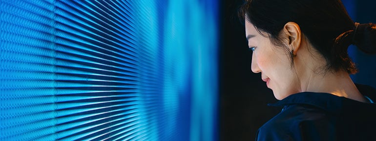 A person holding a mobile phone stands next to a vast digital display wall showing many tones of blue