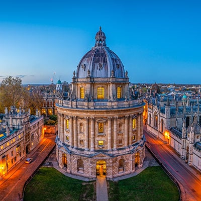 The gothic architecture & cobbled streets of Oxford University glow in warm light at dusk in front of a fading blue sky