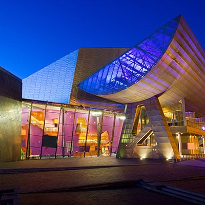The Lowry theatre at night, its metallic surface is lit with purple & blue, through its windows bold tones of purple & orange