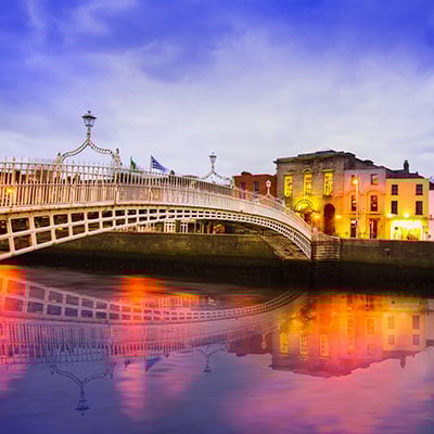Liffey Bridge, Dublin at dusk. Over the river are townhouses & modern glass buildings. The river reflects orange & blue light