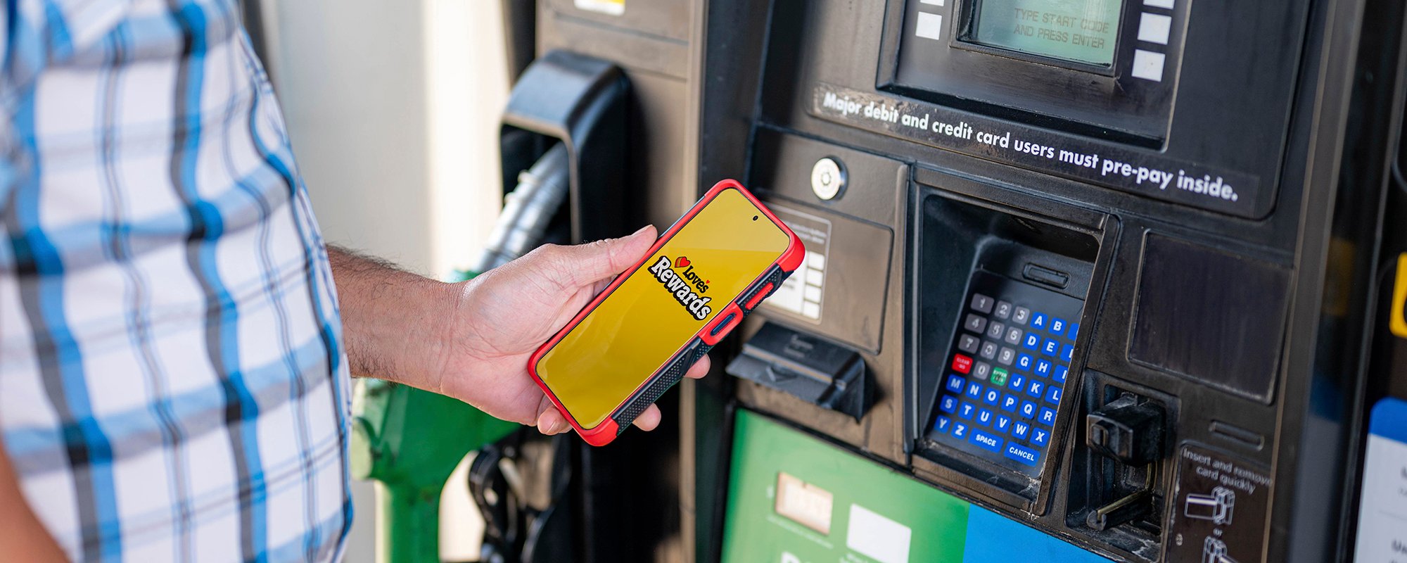 A driver holding a phone at the fuel pump that shows "Love's Rewards" on the homescreen