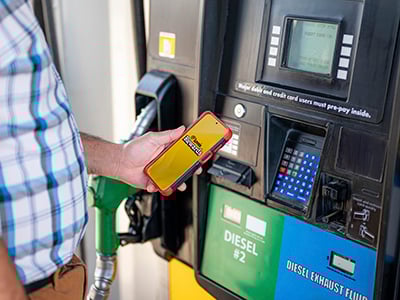 A driver holding a phone at the fuel pump that shows "Love's Rewards" on the homescreen