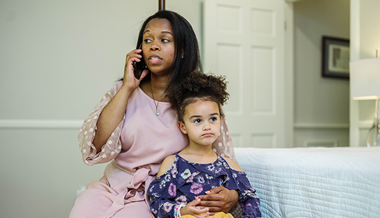 woman in a pink dress talks on the phone while cuddling her young daughter on a bench at the end of a bed
