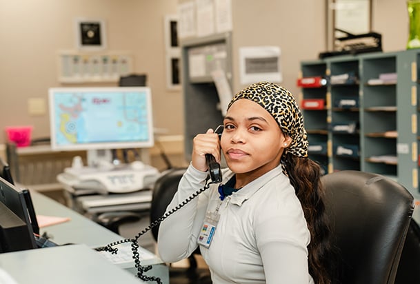 Health care professional wearing a gray longsleeve shirt and leopard head scarf, talking on the phone and looking into the camera