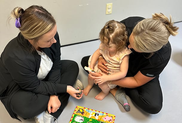 A therapist holds a young girl on her lap while the other therapist hands her puzzle pieces