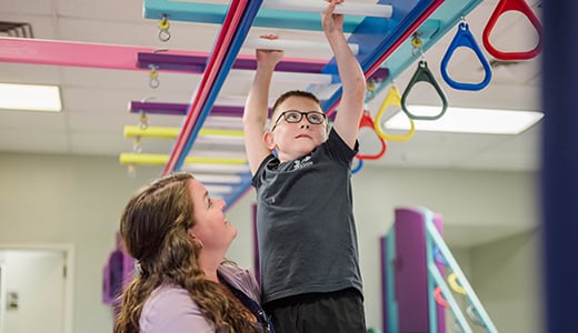 A therapist supports a little boy as he does the monkey bars during therapy