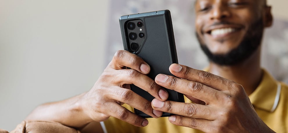 A man in a yellow collared shirt looks into his smartphone while smiling