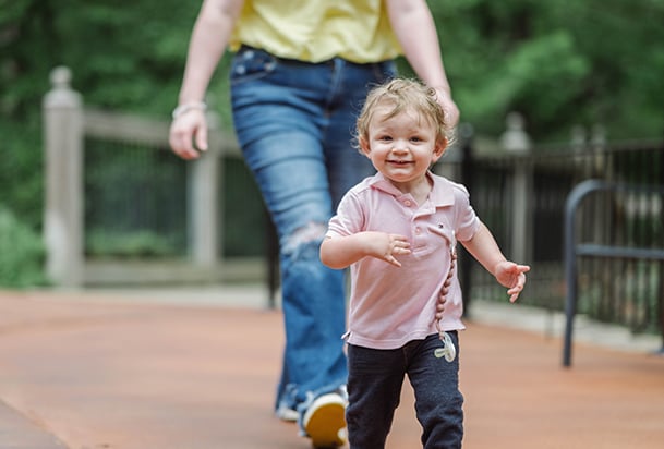 Toddler boy in pink shirt running with mother in yellow shirt following quidkly behind