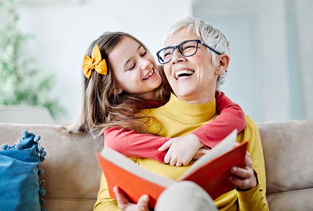 Older woman reading a book laughing while being hugged from behind by a young girl