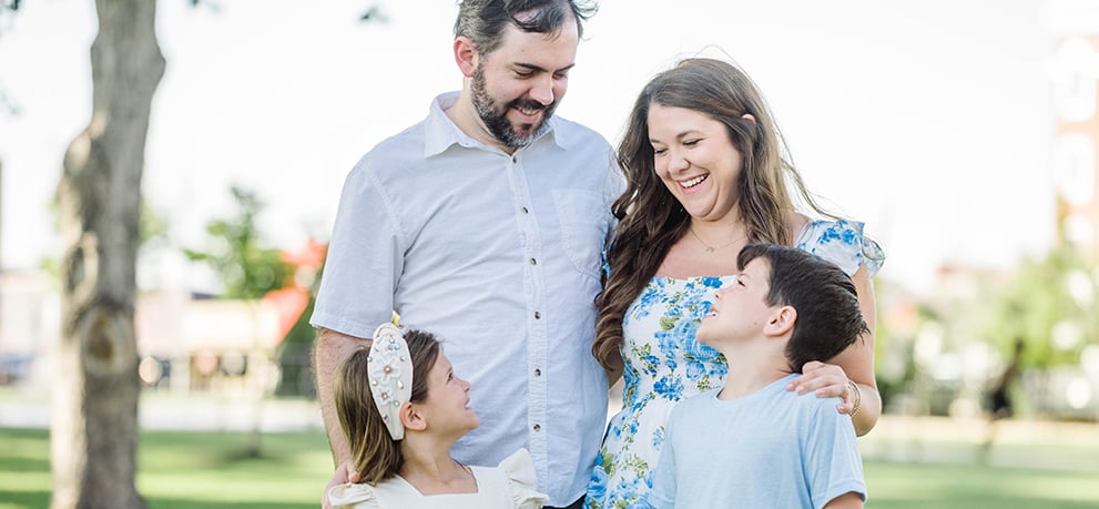 Mother and father smile down at their son and daughter in a park on a sunny day