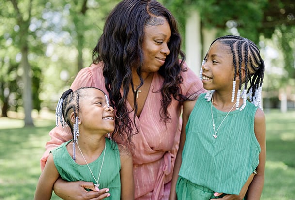 Mother in pink dress hugs her two smiling daughters who are wearing matching green outfits