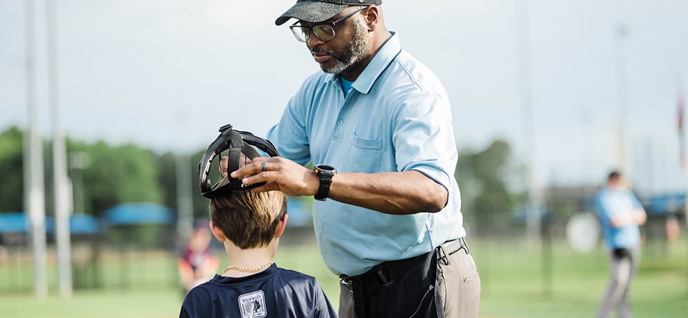 Coach wearing a gray cap and light blue polo shirt puts a helmet on a young boy's head