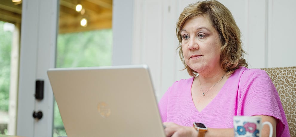 A woman in a pink shirt sitting at a table typing on a laptop with a colorful mug next to her