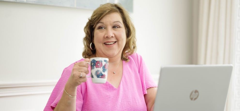 Sitting woman pink top holding coffee cup with laptop on table in front of her