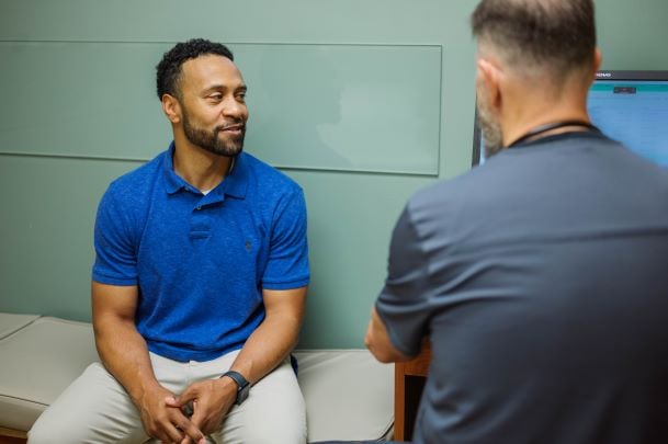 Man sitting on exam table blue top white pants talking to man with stethescope