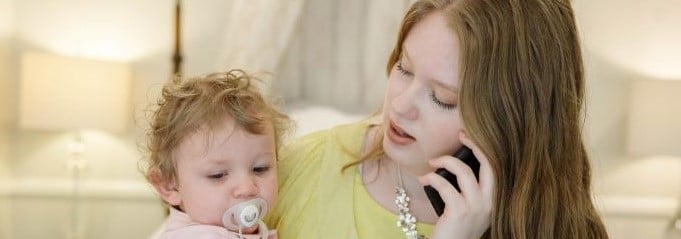 Young mother in yellow top holding baby in light pink, talking on phone