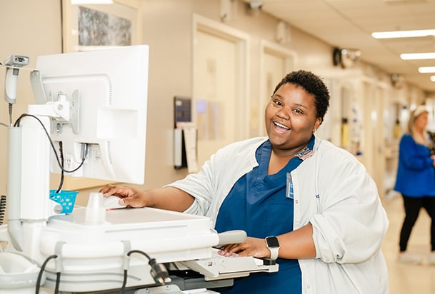 A nurse in blue scrubs and white jacket in front of a computer smiling at the camera