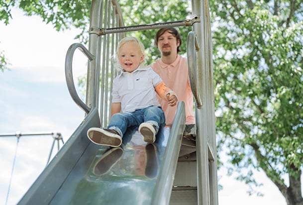 Toddler boy at the top of a slide with his father behind him