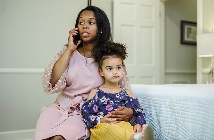 woman in a pink dress talks on the phone while cuddling her young daughter on a bench at the end of a bed