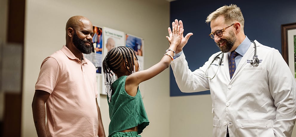 Doctor in a lab coat and little girl in a green outfit sharing a high-five while girl's father looks on