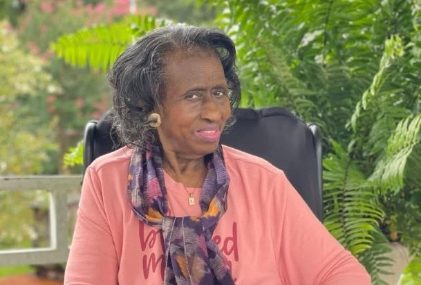 A nursing home resident with a coral outfit and scarf sits outside on a porch