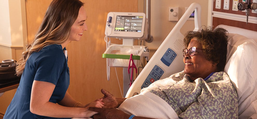 A nurse in dark blue scrubs holds the hands of a smiling patient in a hospital bed