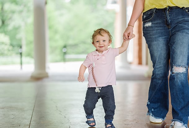 A toddler in a pink top walks while holding his mother's hand