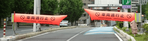 「車両通行止」の文字と進入禁止の標識が黒く描かれた、オレンジ色の巨大なバルーン状の遮断機が道路の両脇から伸びて入口を塞いでいる様子。