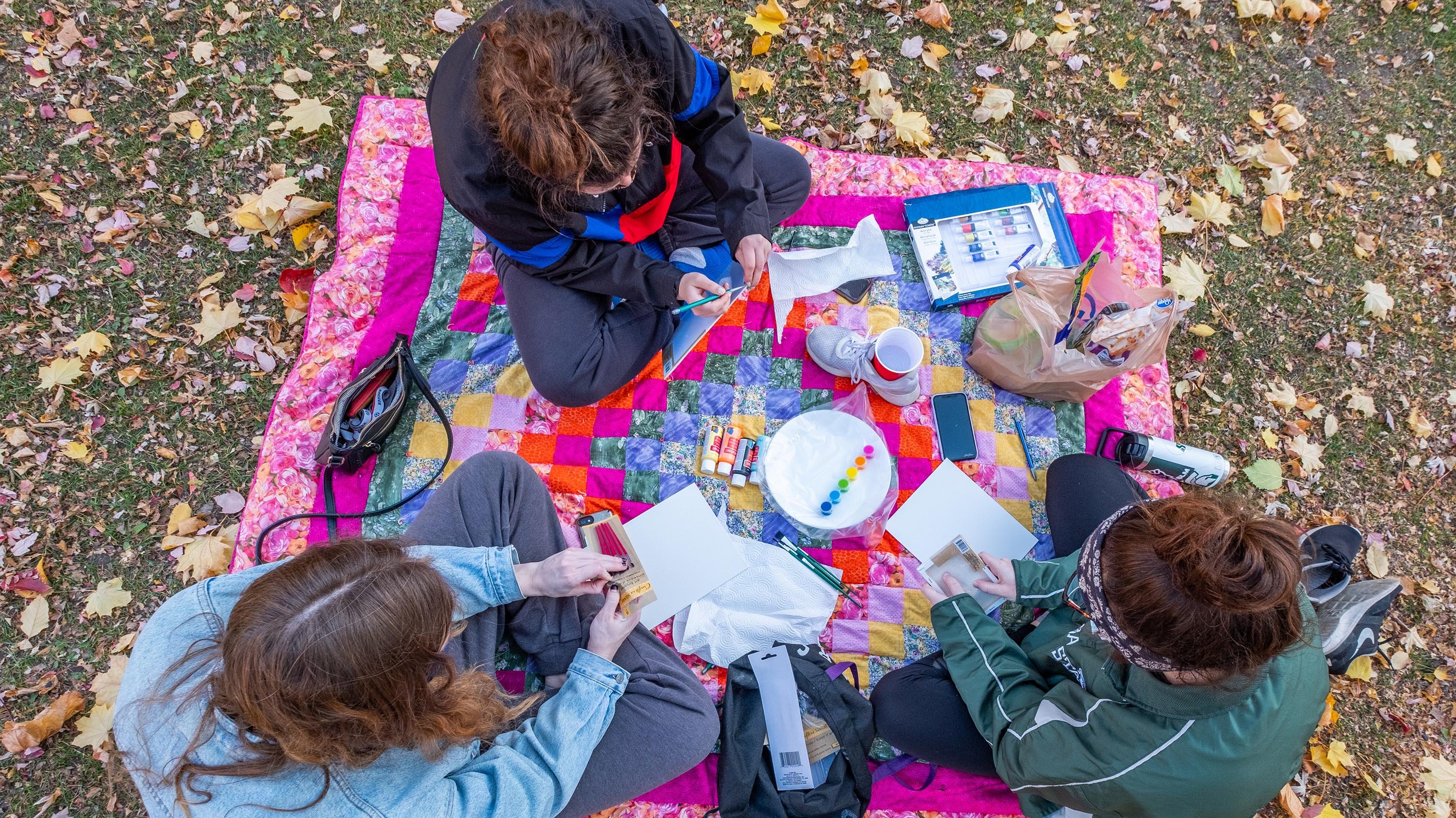 Three students with art supplies, sitting on a blanket in the grass