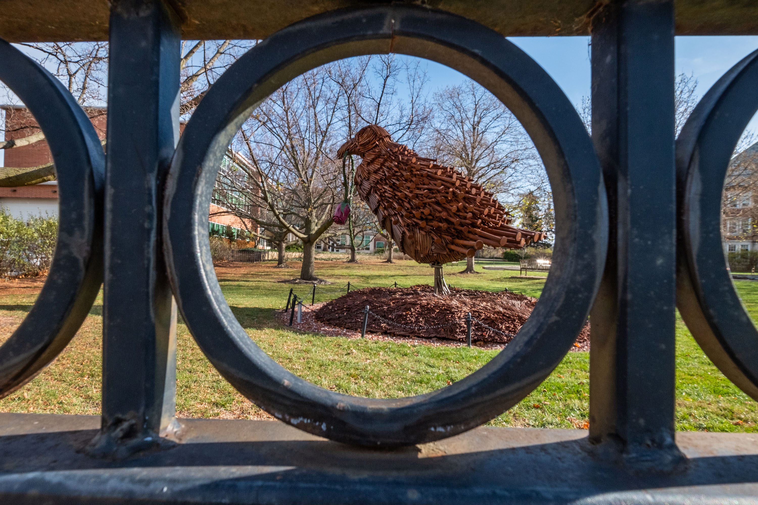 Large sculpture of a bird holding a rose in its mouth as seen through a circular fence opening